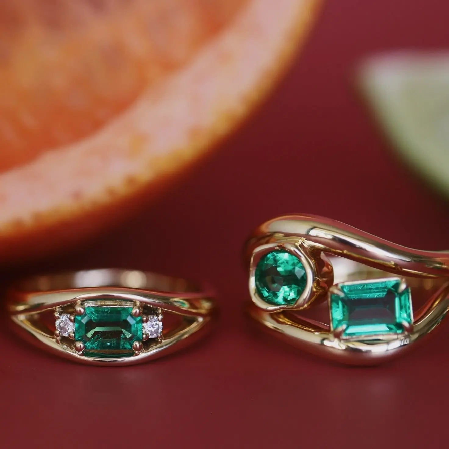 Two gold rings with emerald stones on a red surface.