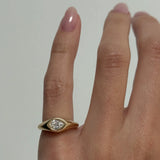 A close-up of a hand wearing a yellow gold signet ring with a pear-cut diamond in a bezel setting, against a plain light gray background.