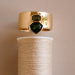 Gold ring with green gemstones on a spool of thread against a beige background