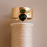 Gold ring with green gemstones on a spool of thread against a beige background