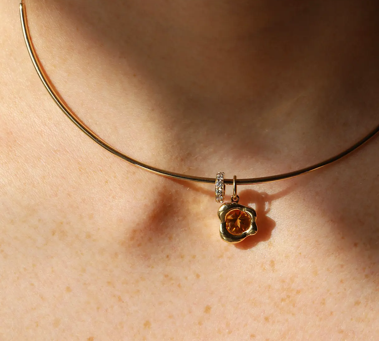 A close-up of a person's neck wearing a delicate gold collar necklace with a small abstract bezel gold pendant featuring a orange citrine stone and a small diamond rondelle.
