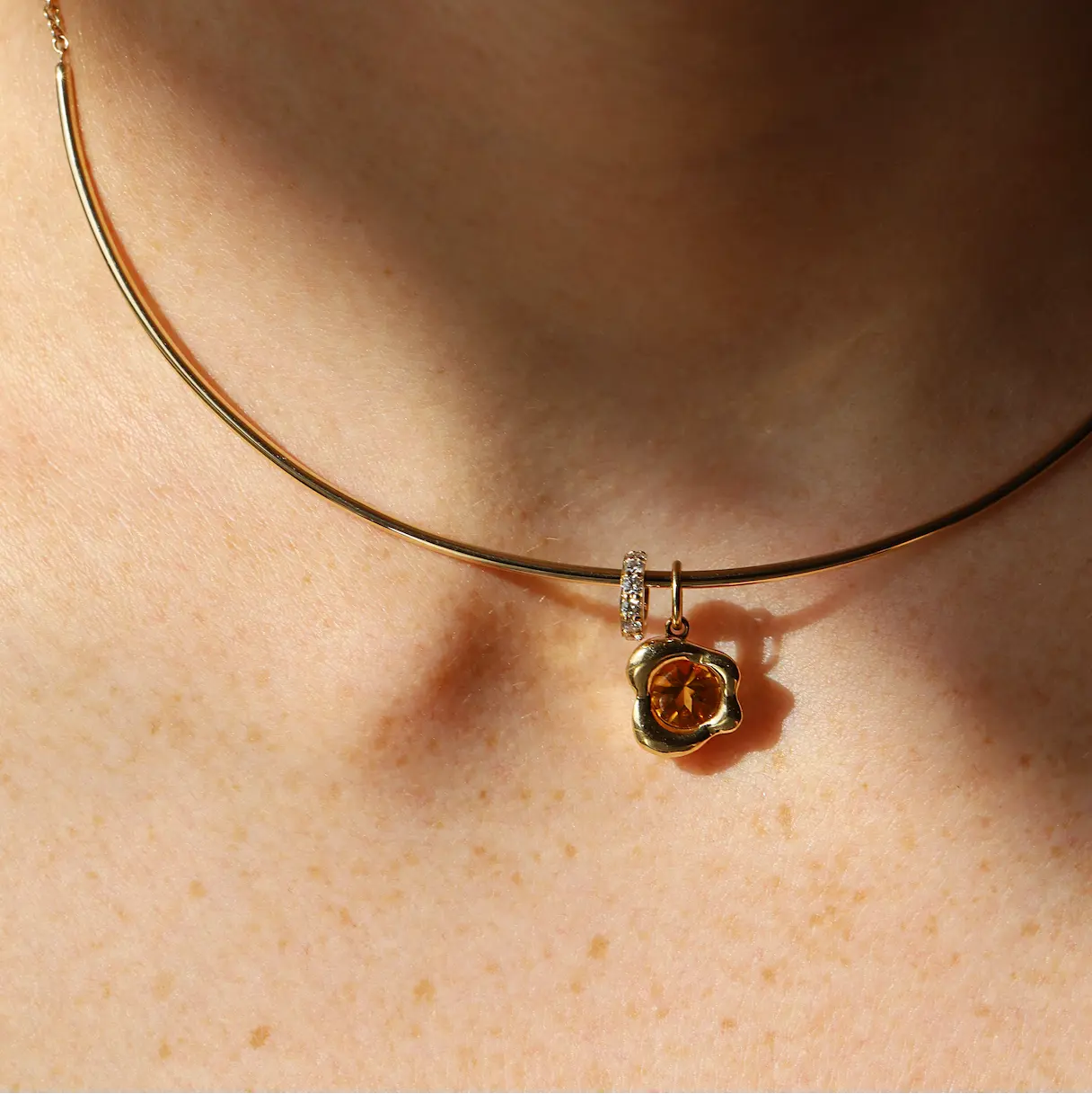 A close-up of a person's neck wearing a delicate gold collar necklace with a small abstract bezel gold pendant featuring a orange citrine stone and a small diamond rondelle.