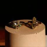 Two gold rings with gemstones on a cylindrical stand against a dark background