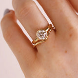 A close-up of a hand wearing a gold ring with a round clear gemstone in a prong setting, photographed against a soft light background.