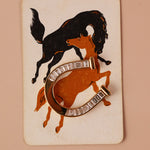 A vintage-style horseshoe-shaped brooch with clear rectangular stones, overlaid on an orange and black illustration of a horse and rider on a cream card, set against a solid peach background.