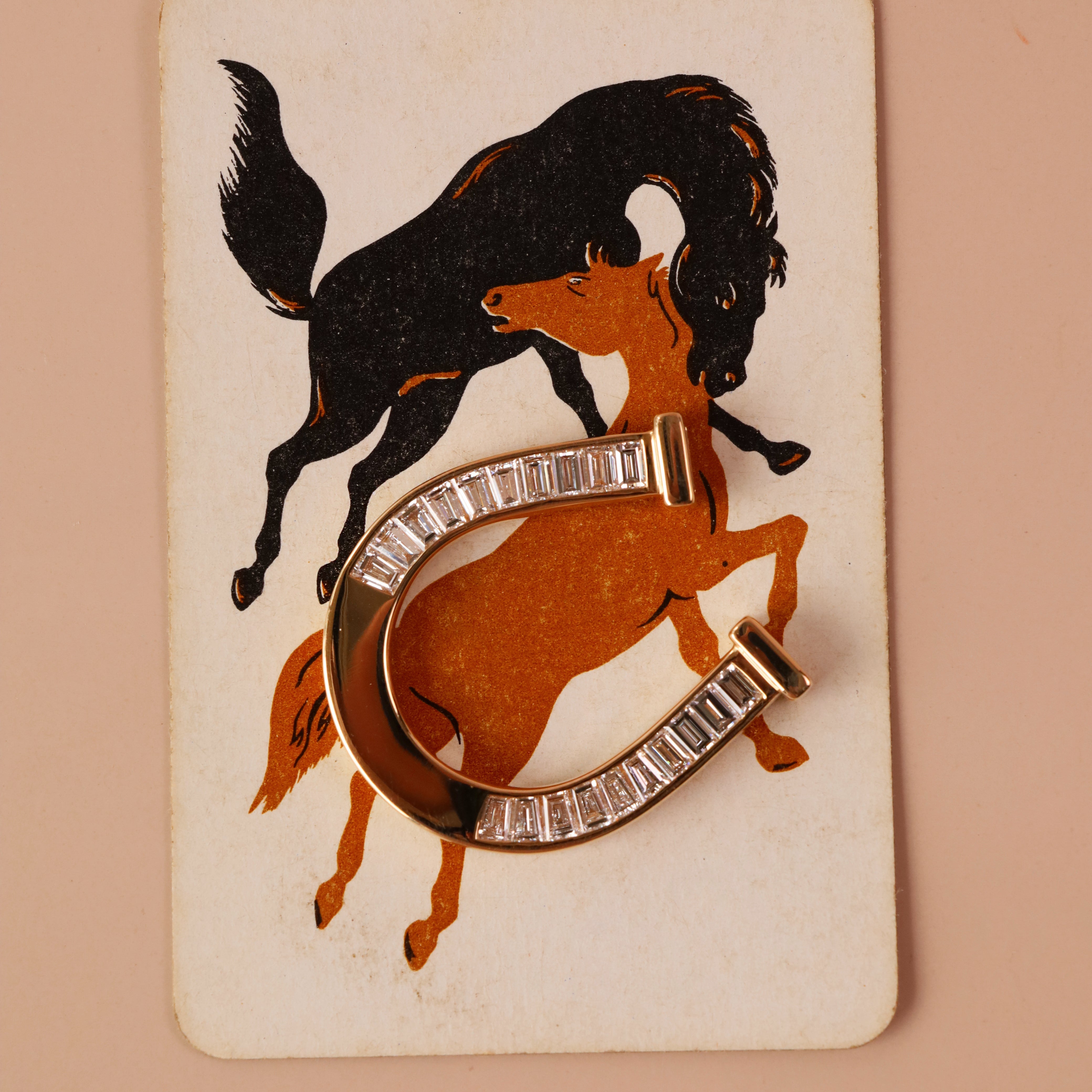 A vintage-style horseshoe-shaped brooch with clear rectangular stones, overlaid on an orange and black illustration of a horse and rider on a cream card, set against a solid peach background.