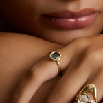 Close-up of a person's hand and lower face wearing multiple gold rings. One ring features a dark blue-gray stone in a hexagonal bezel, while another larger ring has an oval clear stone with small side stones and sculptural gold bands.