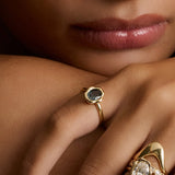 Close-up of a person's hand and lower face wearing multiple gold rings. One ring features a dark blue-gray stone in a hexagonal bezel, while another larger ring has an oval clear stone with small side stones and sculptural gold bands.