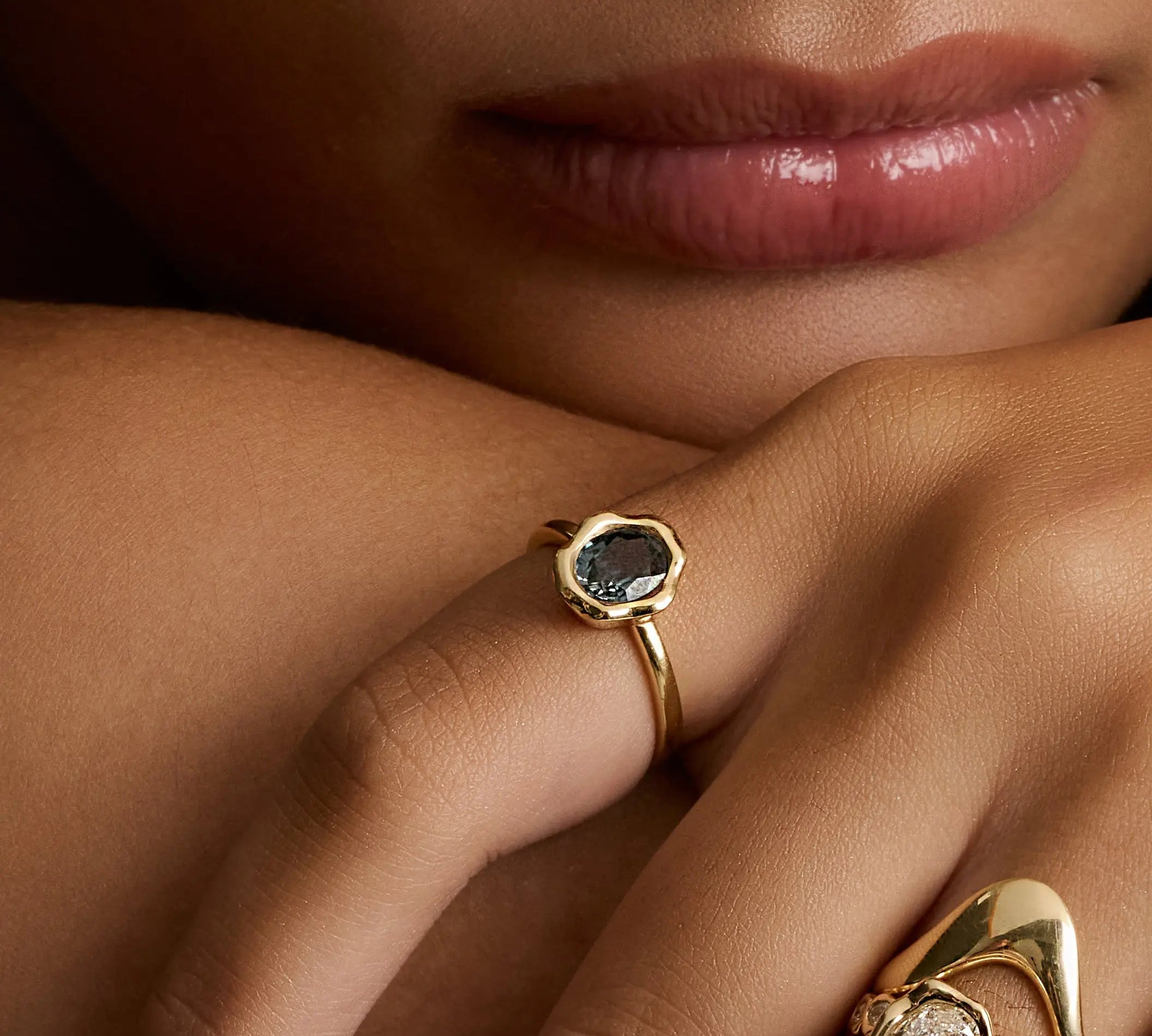 Close-up of a person's hand and lower face wearing multiple gold rings. One ring features a dark blue-gray stone in a hexagonal bezel, while another larger ring has an oval clear stone with small side stones and sculptural gold bands.