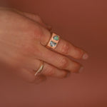 Hand wearing a peach enamel gold ring with gemstones against a warm brown background