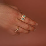 Hand wearing a peach enamel gold ring with gemstones against a warm brown background