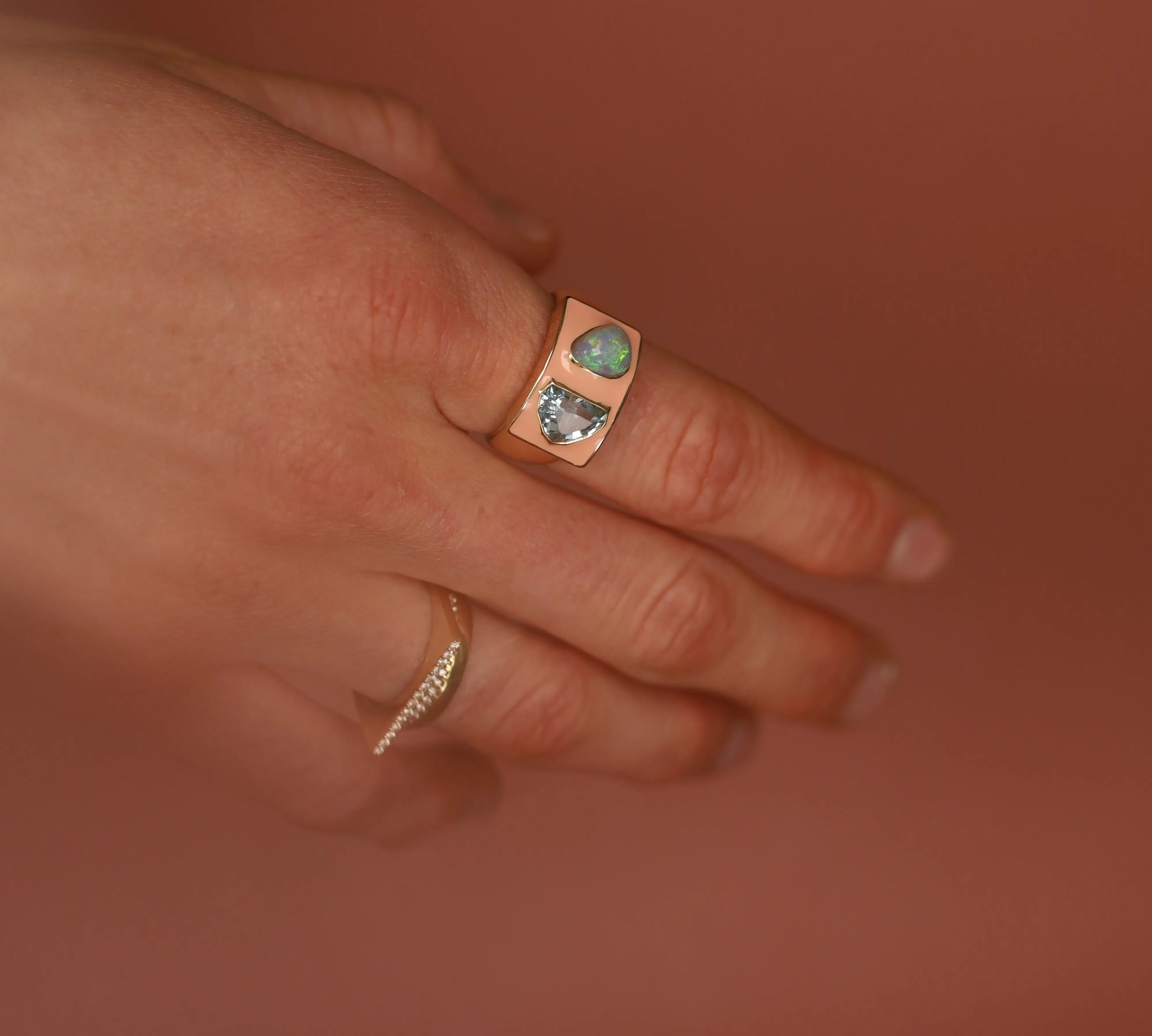Hand wearing a peach enamel gold ring with gemstones against a warm brown background