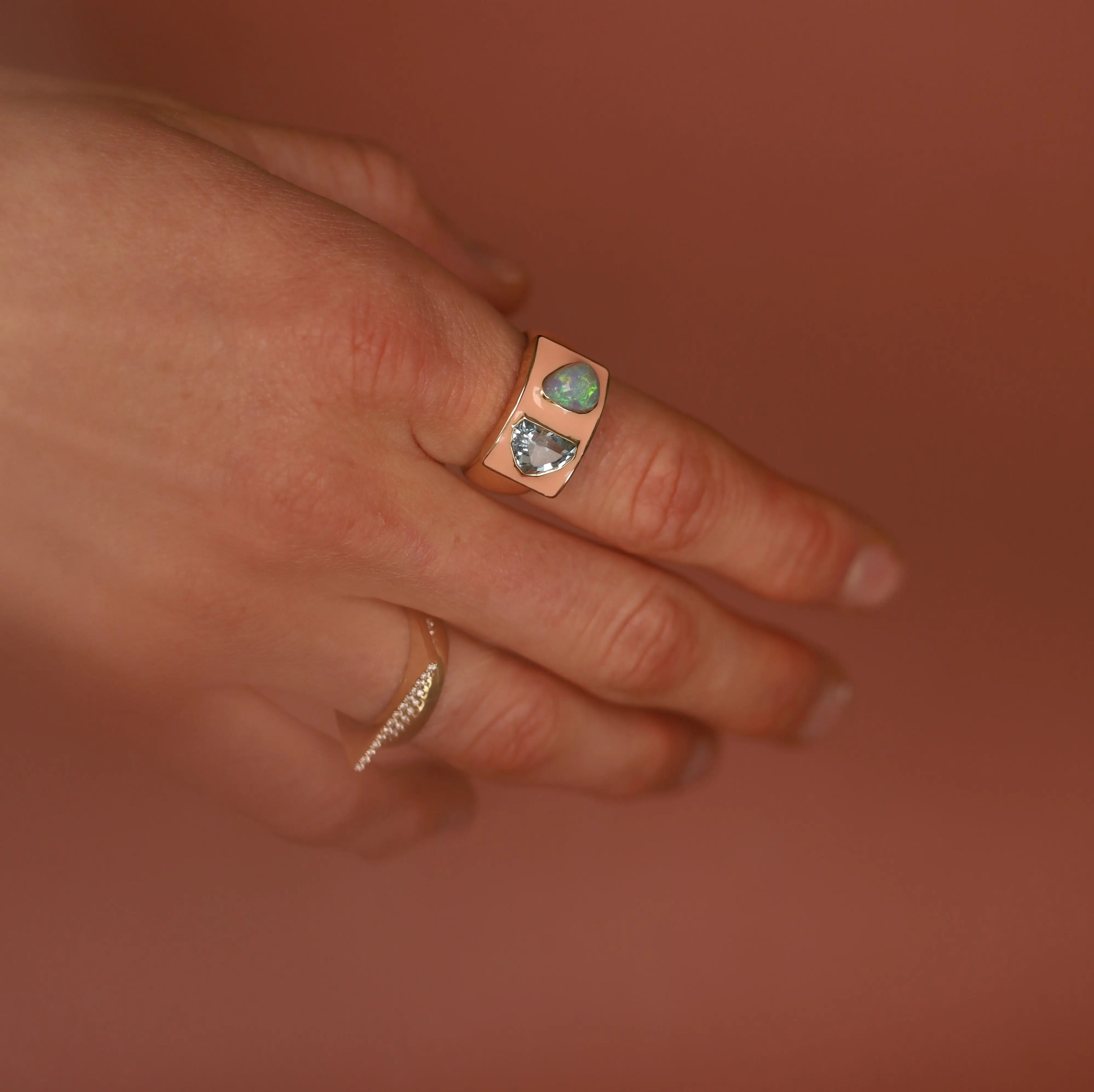 Hand wearing a peach enamel gold ring with gemstones against a warm brown background