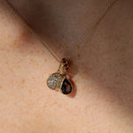 A close-up of a delicate gold chain necklace worn on skin, featuring a pendant with a round pavé-set clear stone and a teardrop-shaped dark smoky gemstone in a gold bezel.