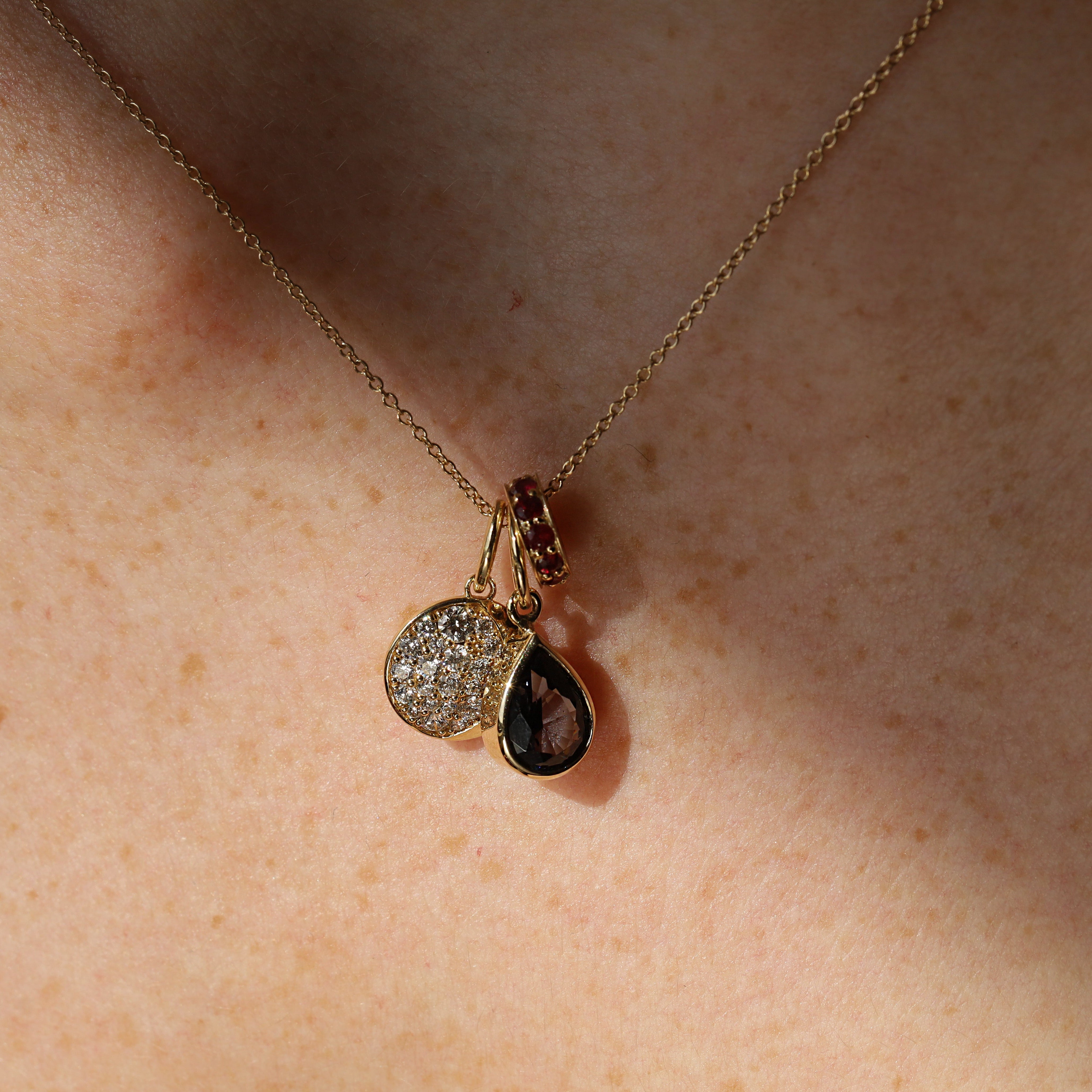A close-up of a delicate gold chain necklace worn on skin, featuring a pendant with a round pavé-set clear stone and a teardrop-shaped dark smoky gemstone in a gold bezel.