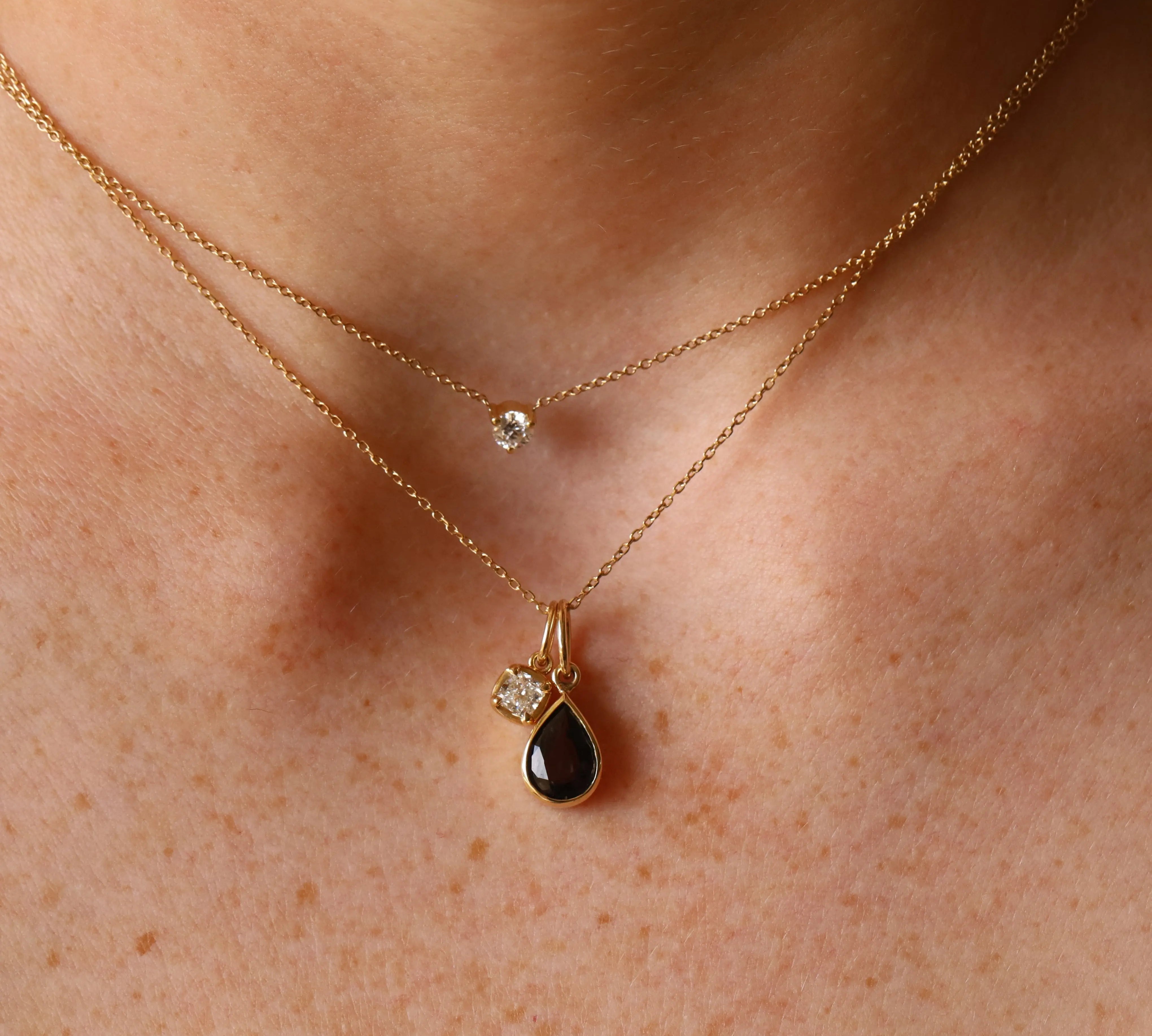 Close up of woman's décolletage wearing a dainty round diamond choker, layered with another dainty gold chain that has a grey spinel pear charm hanging from it, alongside a smaller cushion cut diamond charm.