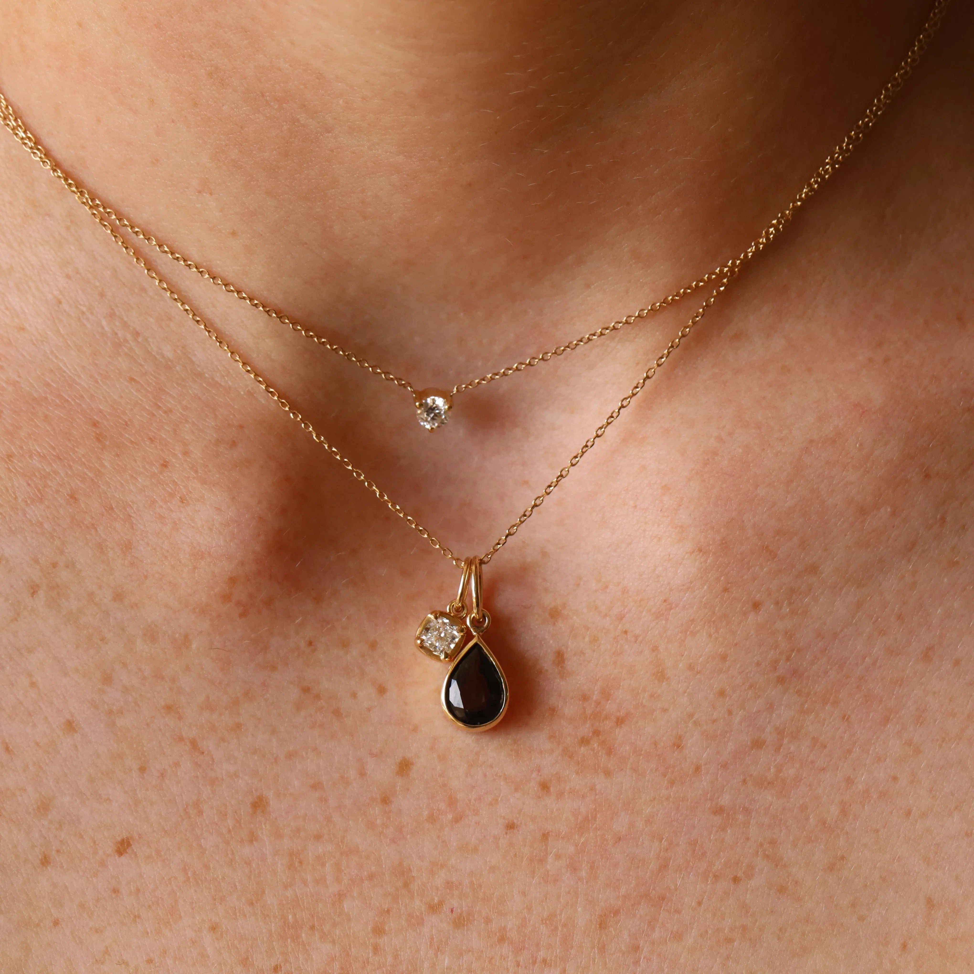 Close up of woman's décolletage wearing a dainty round diamond choker, layered with another dainty gold chain that has a grey spinel pear charm hanging from it, alongside a smaller cushion cut diamond charm.