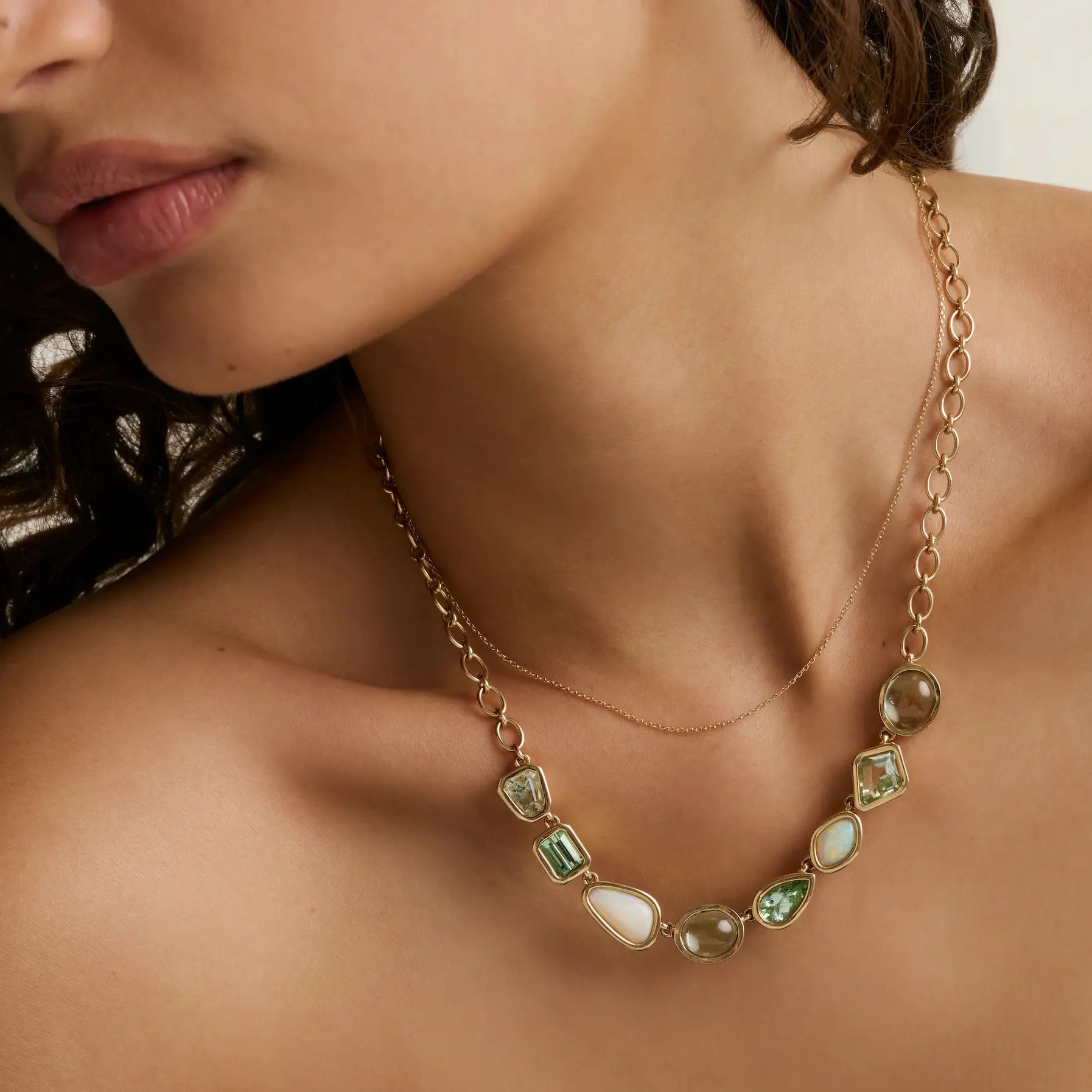 close up of woman's decolletage wearing a dainty yellow gold chain layered with a chunky necklace with green colored stones in different shapes and sizes