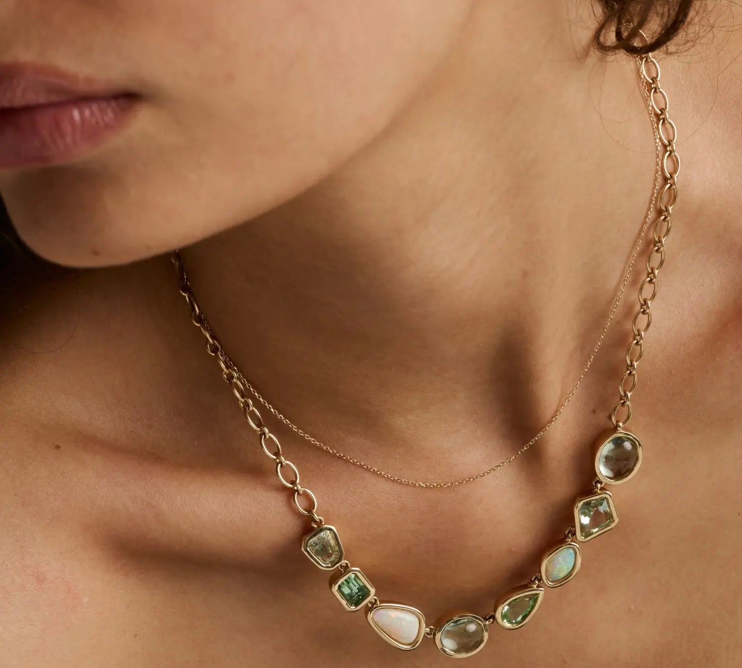 A close-up of a model wearing a gold necklace with irregular green and white gemstones, layered over a delicate gold chain.