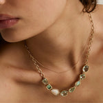 A close-up of a model wearing a gold necklace with irregular green and white gemstones, layered over a delicate gold chain.