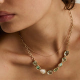 A close-up of a model wearing a gold necklace with irregular green and white gemstones, layered over a delicate gold chain.