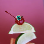 Cherry with a green gemstone on a lime wedge with a blurred orange slice below on a pink background