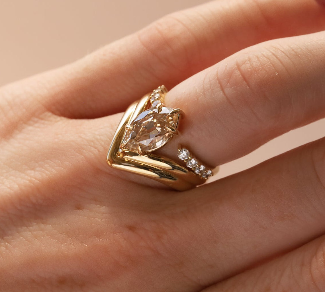 A gold ring with a marquise-cut clear center stone stacked with open shank white diamond band, worn on a person's finger against a soft beige background.