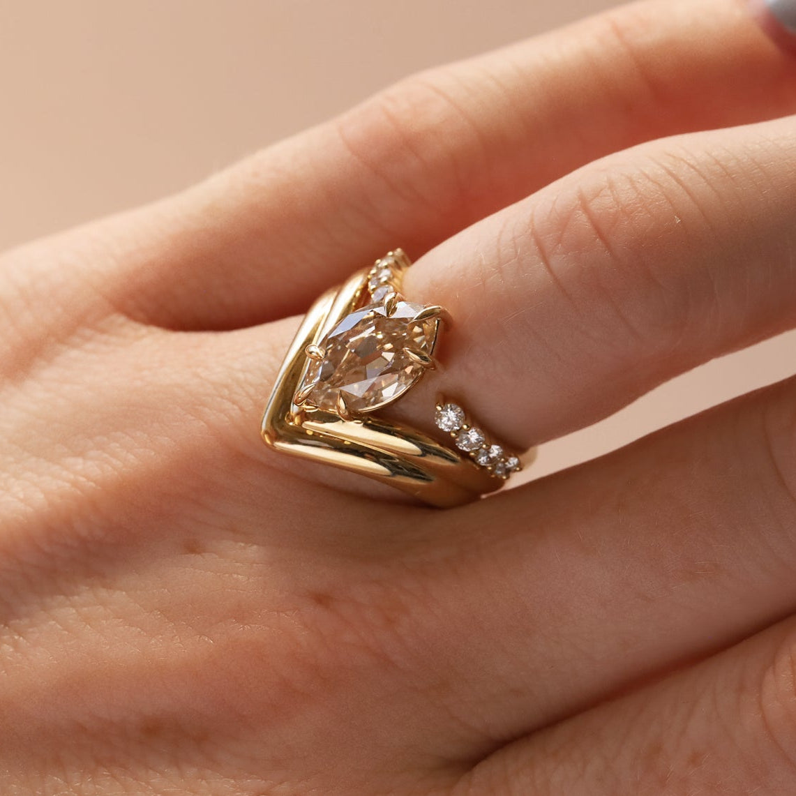 A gold ring with a marquise-cut clear center stone stacked with open shank white diamond band, worn on a person's finger against a soft beige background.