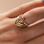 A gold ring with a marquise-cut clear center stone stacked with open shank white diamond band, worn on a person's finger against a soft beige background.
