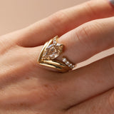 A gold ring with a marquise-cut clear center stone stacked with open shank white diamond band, worn on a person's finger against a soft beige background.