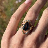Hand wearing a gold ring with a black gemstone against a blurred natural background
