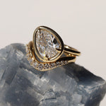 Tilted pear diamond ring sitting on top of a white diamond wave stacking band. Both on top of a grey-blue gemstone.