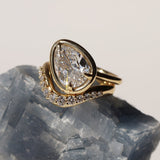 Tilted pear diamond ring sitting on top of a white diamond wave stacking band. Both on top of a grey-blue gemstone.