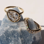 Tilted pear diamond ring and white diamond wave stacking band sitting on top of a grey-blue gemstone.