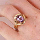 A gold ring with a large lavender-purple oval gemstone, worn on a person's finger against a soft pink background.