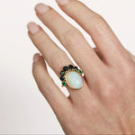 Hand wearing a ring with an opal stone and green gemstones on a plain background