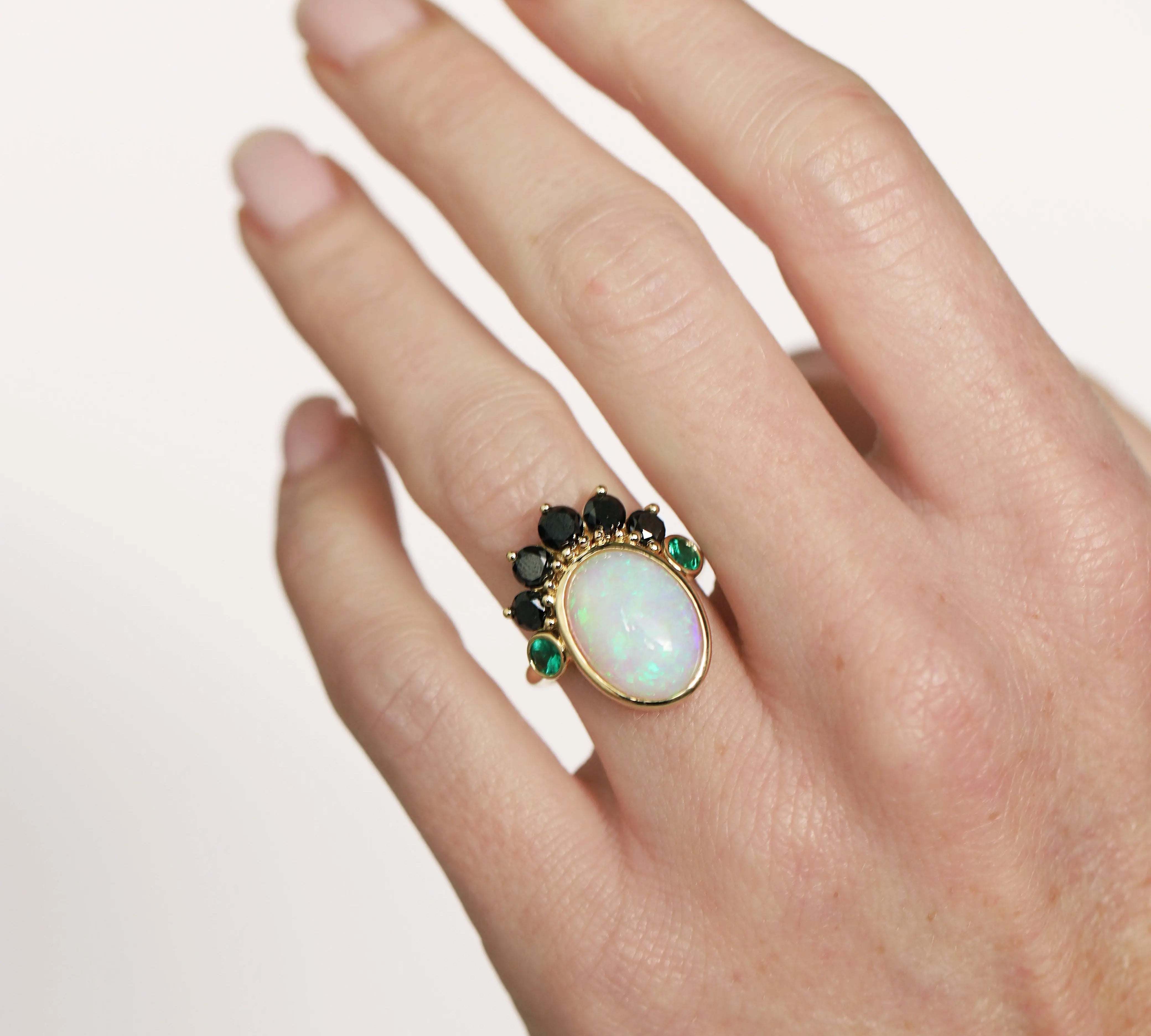 Hand wearing a ring with an opal stone and green gemstones on a plain background