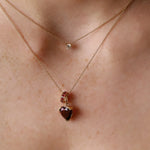 A close-up of a person's neck wearing two delicate gold necklaces. The shorter necklace has a small clear round stone, while the longer necklace features a heart-shaped faceted gemstone pendant with a small gold ring detail above it.