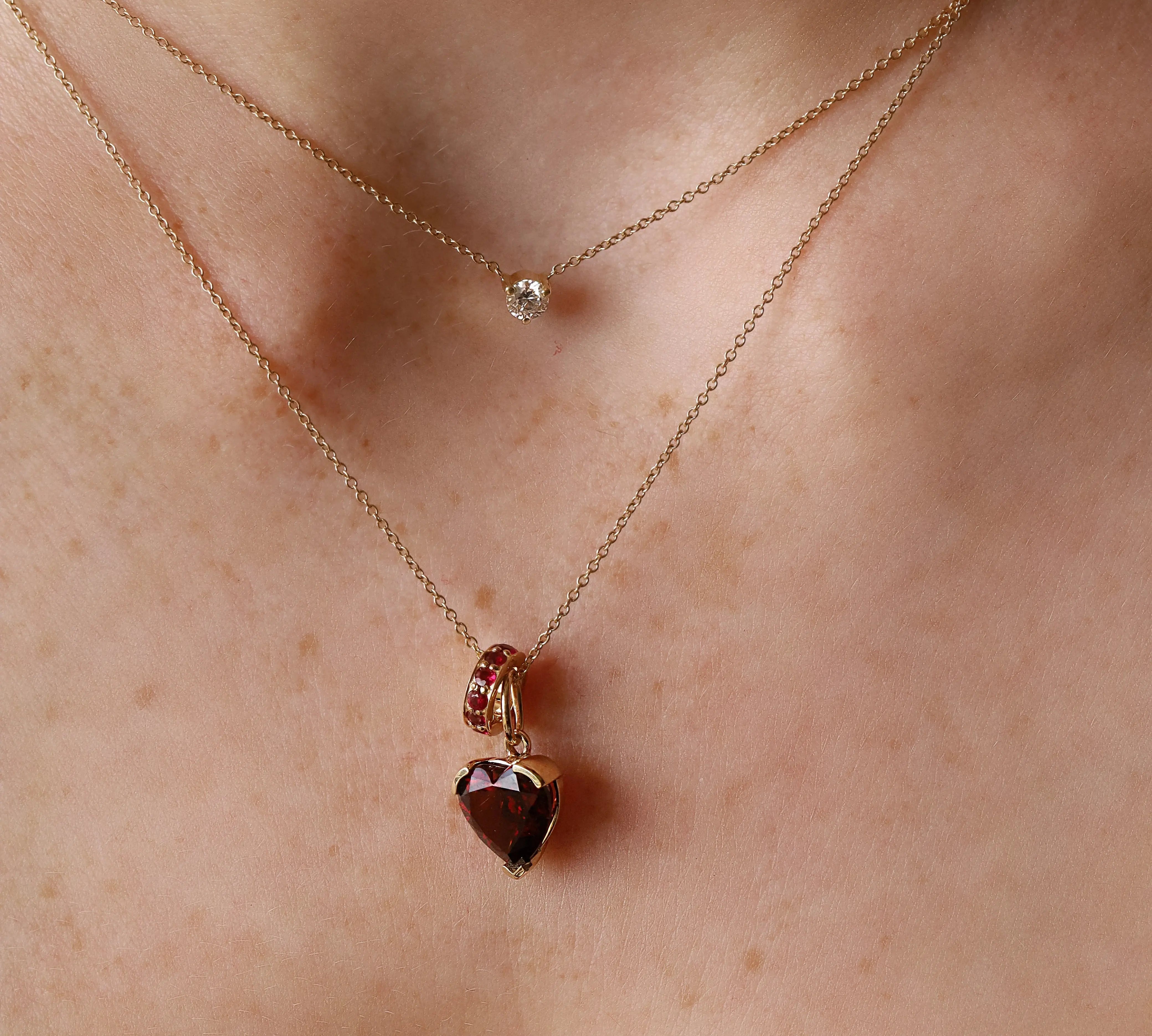 A close-up of a person's neck wearing two delicate gold necklaces. The shorter necklace has a small clear round stone, while the longer necklace features a heart-shaped faceted gemstone pendant with a small gold ring detail above it.