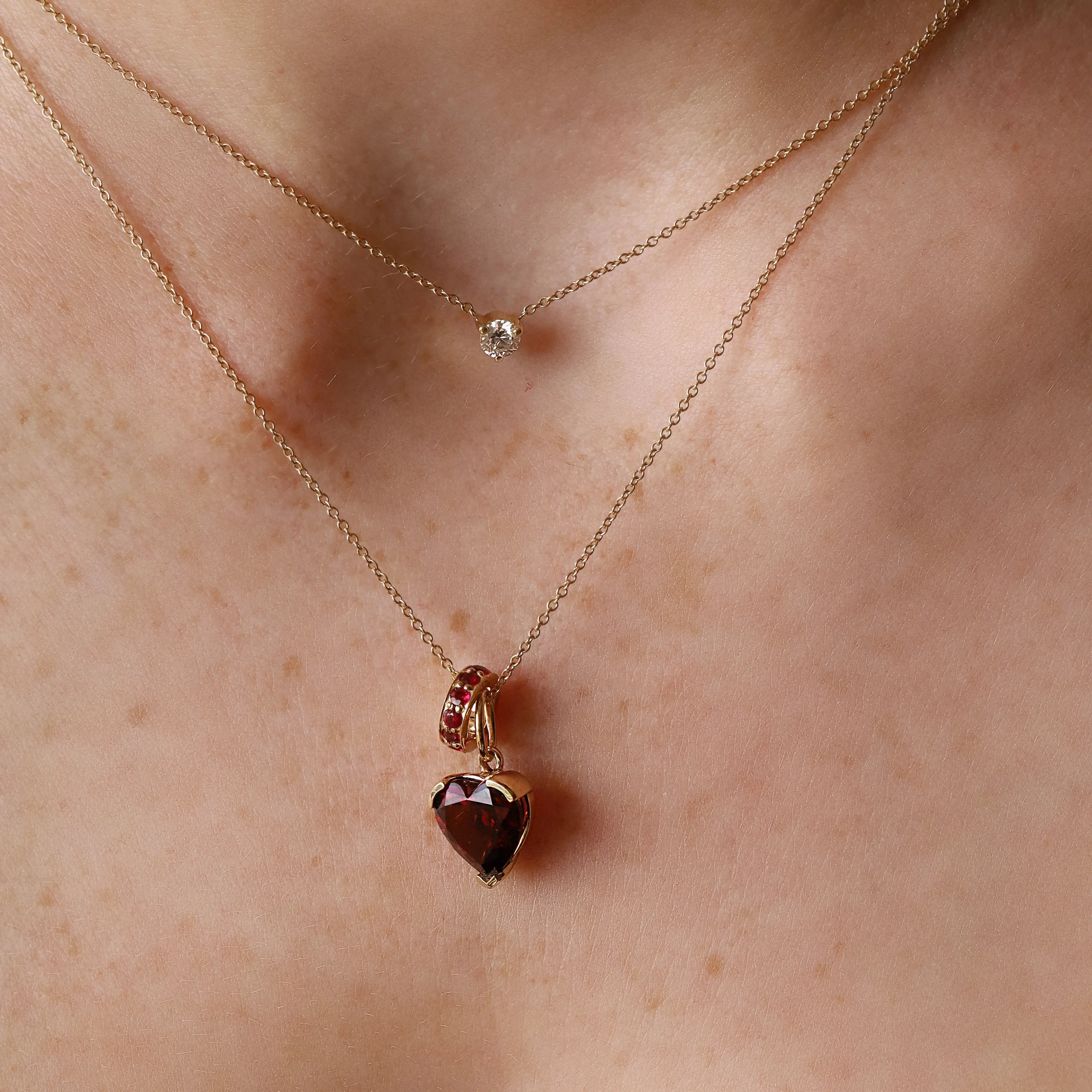 A close-up of a person's neck wearing two delicate gold necklaces. The shorter necklace has a small clear round stone, while the longer necklace features a heart-shaped faceted gemstone pendant with a small gold ring detail above it.