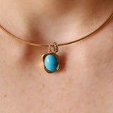 A close-up of a gold bangle necklace with a turquoise cabochon pendant, worn on a person's neck.
