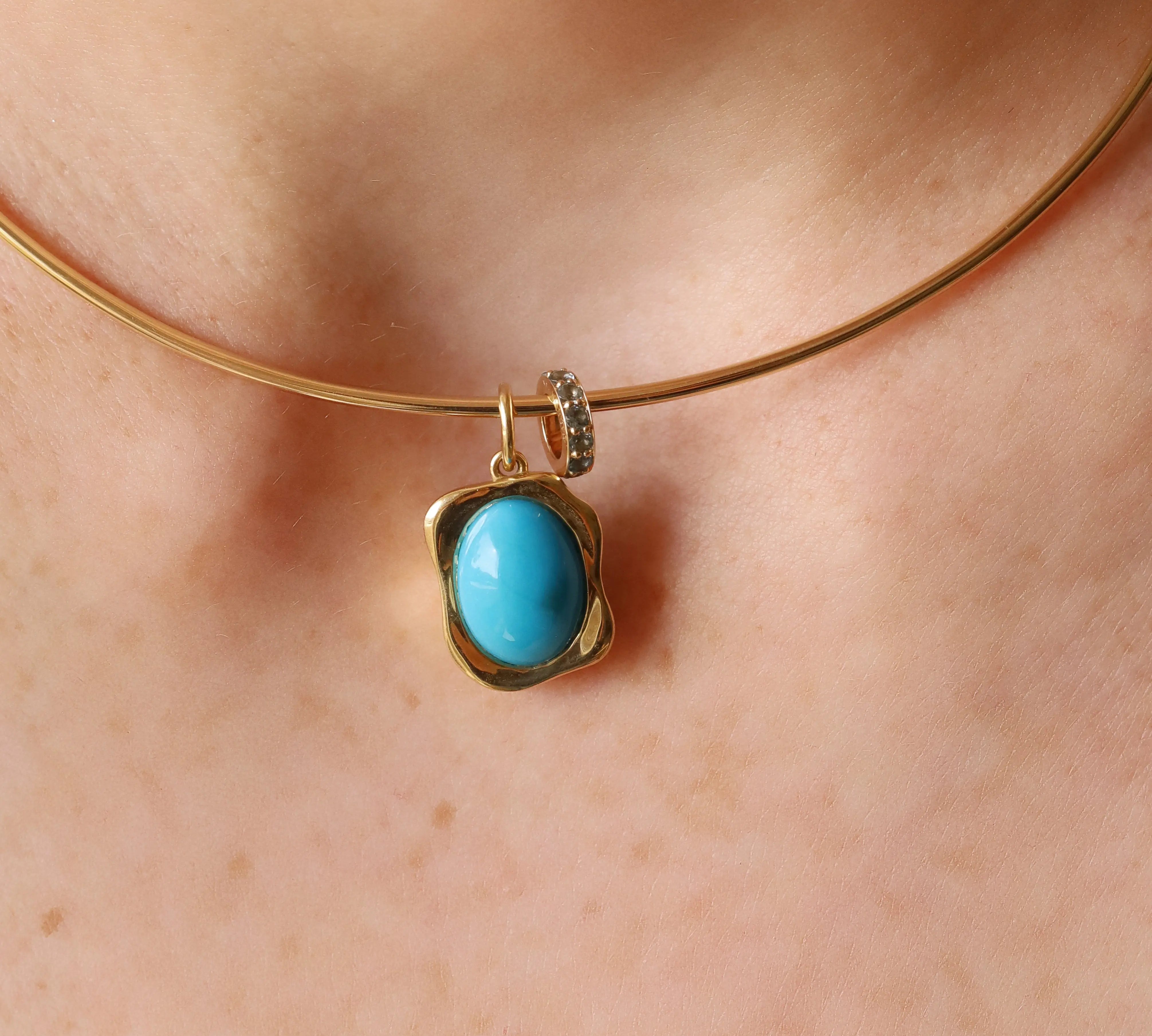 A close-up of a gold bangle necklace with a turquoise cabochon pendant, worn on a person's neck.