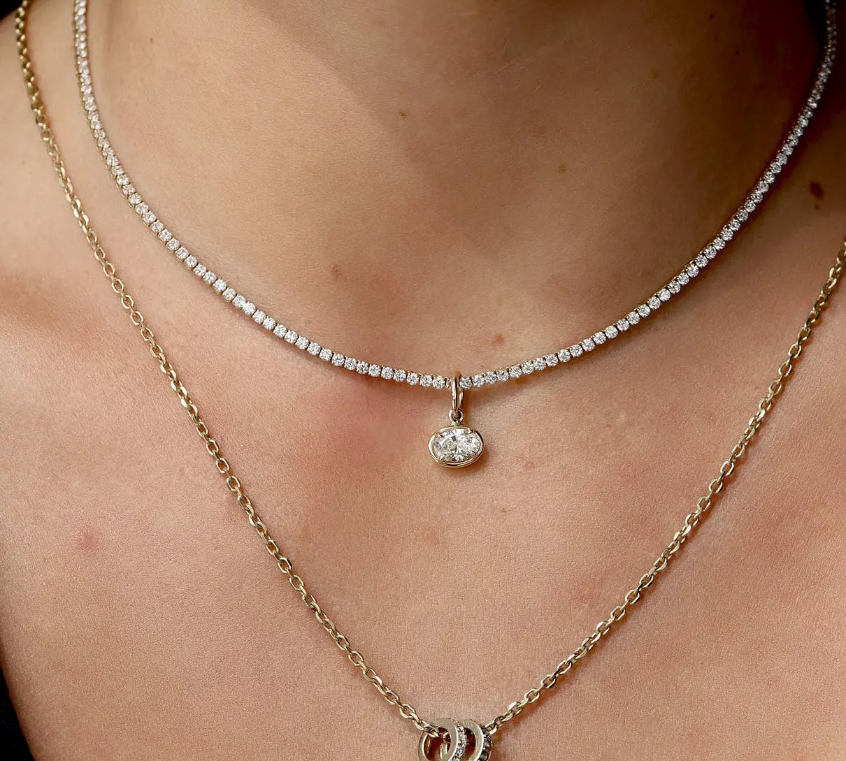 Close-up of model's neck with two necklaces. One tennis necklace with a diamond charm on it, and one gold chain with two rondelle charms