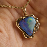 A gold pendant necklace with a heart-shaped iridescent opal stone, held in a person's hand. The pendant features a wavy gold border with small clear accent stones on the right side.
