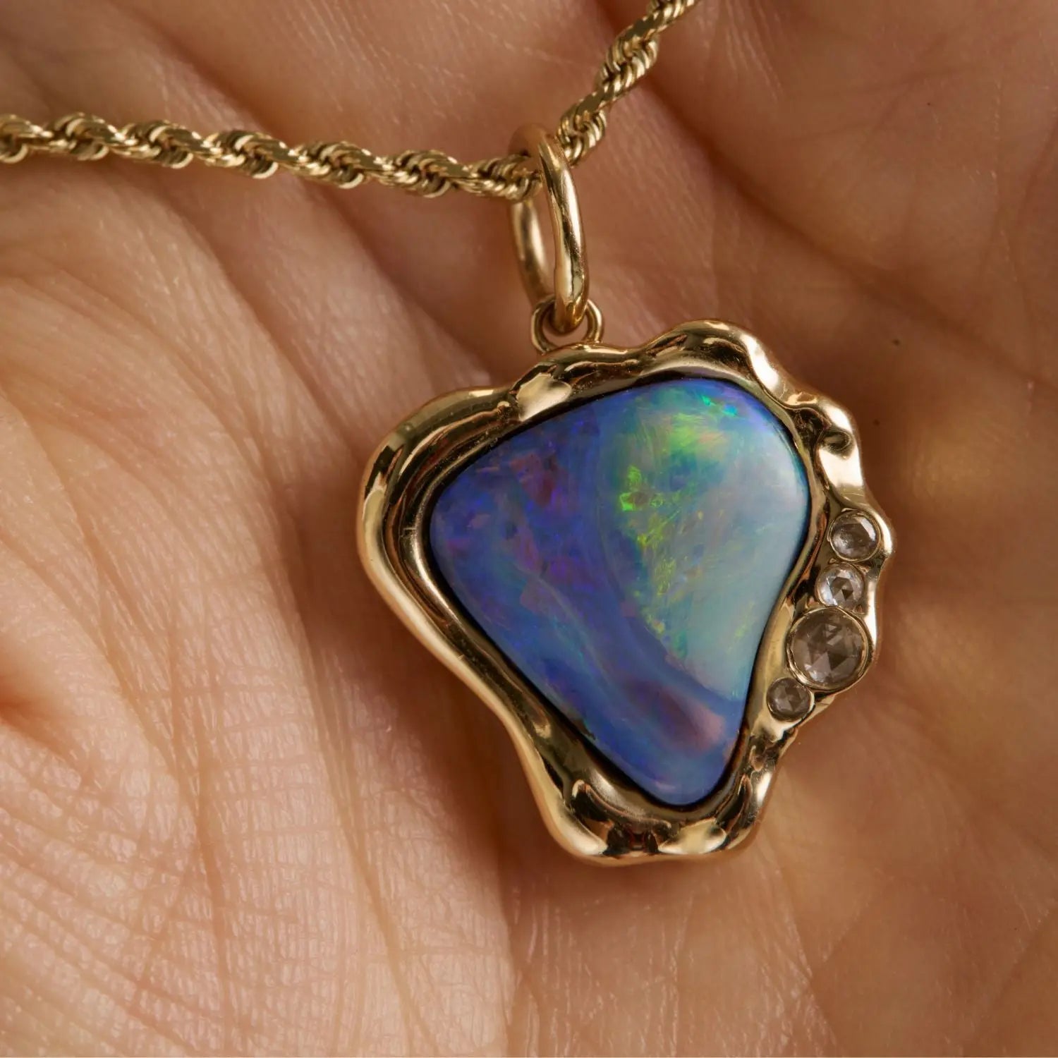A gold pendant necklace with a heart-shaped iridescent opal stone, held in a person's hand. The pendant features a wavy gold border with small clear accent stones on the right side.
