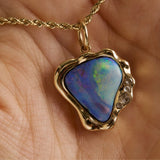 A gold pendant necklace with a heart-shaped iridescent opal stone, held in a person's hand. The pendant features a wavy gold border with small clear accent stones on the right side.