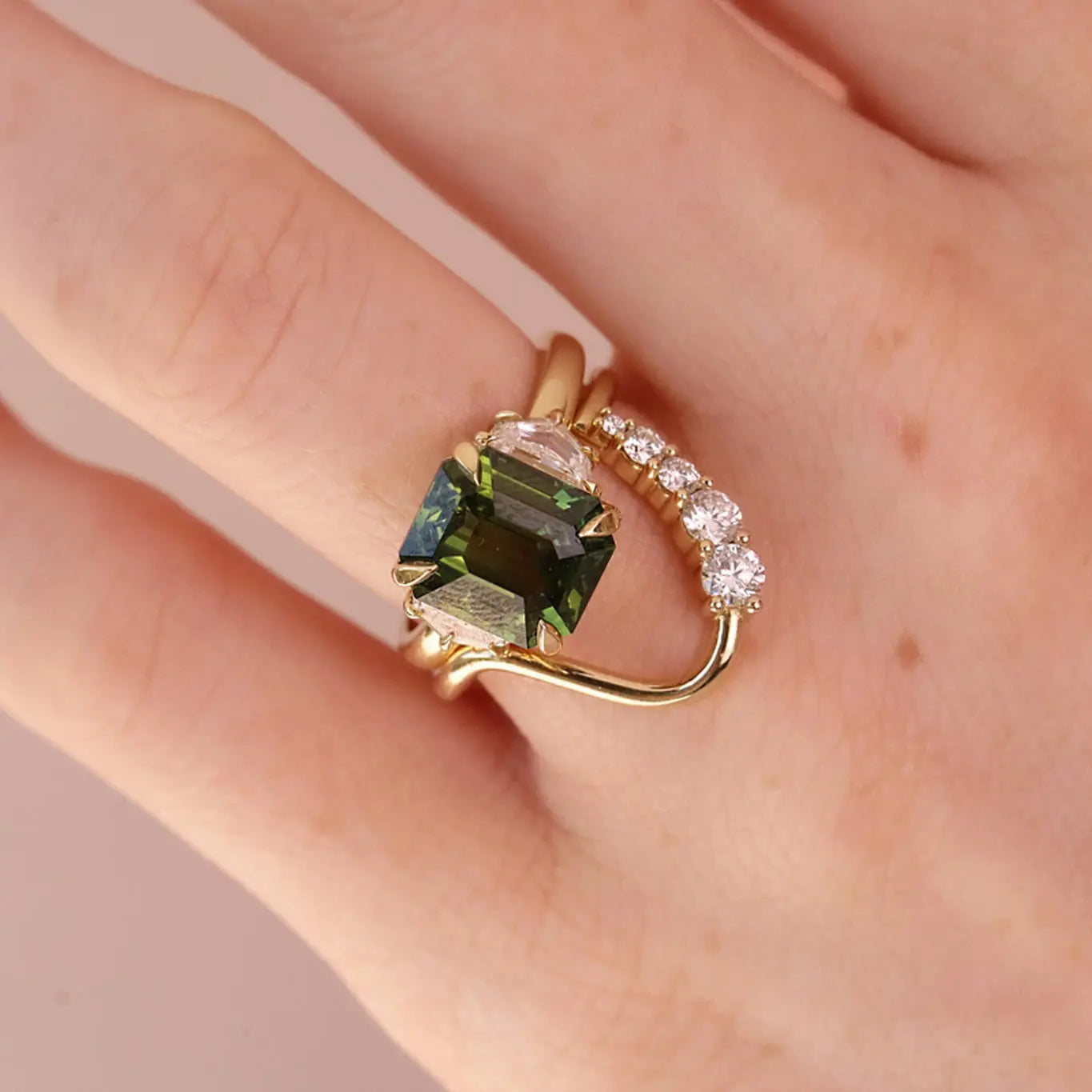 A gold ring with a large emerald-cut green gemstone and a curved pavé band with clear stones, shown worn on a hand against a soft pink background.