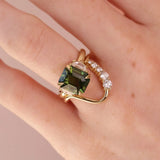 A gold ring with a large emerald-cut green gemstone and a curved pavé band with clear stones, shown worn on a hand against a soft pink background.