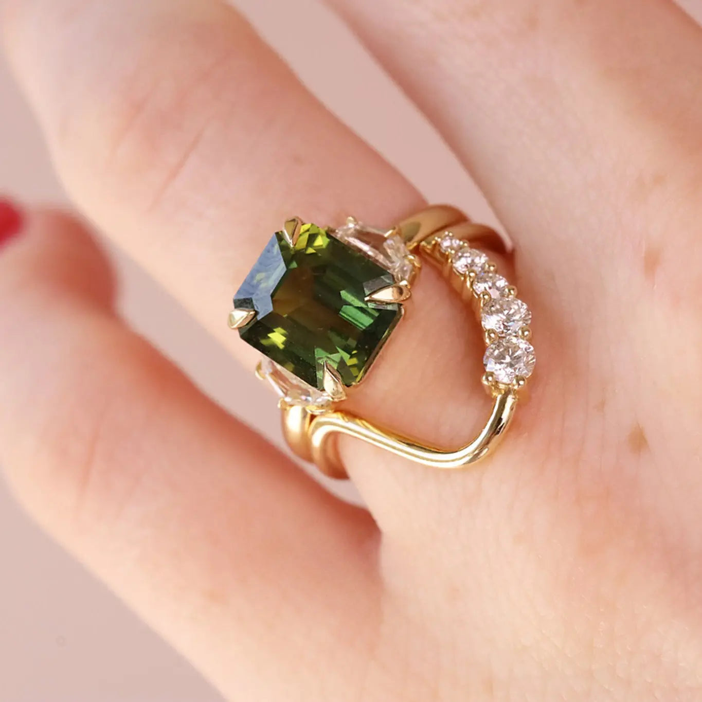 A gold ring with a large emerald-cut green gemstone and a curved band set with small clear stones, shown worn on a hand.