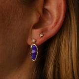 Close-up of an ear with a purple gemstone earring and gold stud.